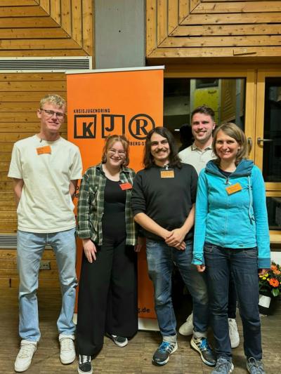 KJR-VV Frühjahr 2025 BDKJ Delegierte (c) BDKJ Fünf BDKJ-Delegierte stehen lächelnd vor einem orangefarbenen KJR-Banner bei der KJR Vollversammlung in einem Raum mit Holzverkleidung. Die Delegierten sind von links nach rechts: Moritz Schuka, Sophie Will, Andreas Löw, Lukas Hochgesang und Melanie Zink.
