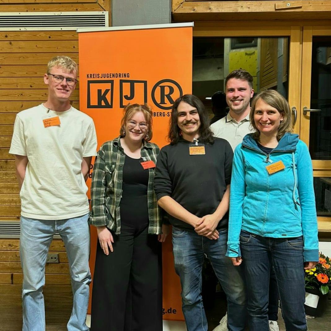 Fünf BDKJ-Delegierte stehen lächelnd vor einem orangefarbenen KJR-Banner bei der KJR Vollversammlung in einem Raum mit Holzverkleidung. Die Delegierten sind von links nach rechts: Moritz Schuka, Sophie Will, Andreas Löw, Lukas Hochgesang und Melanie Zink.