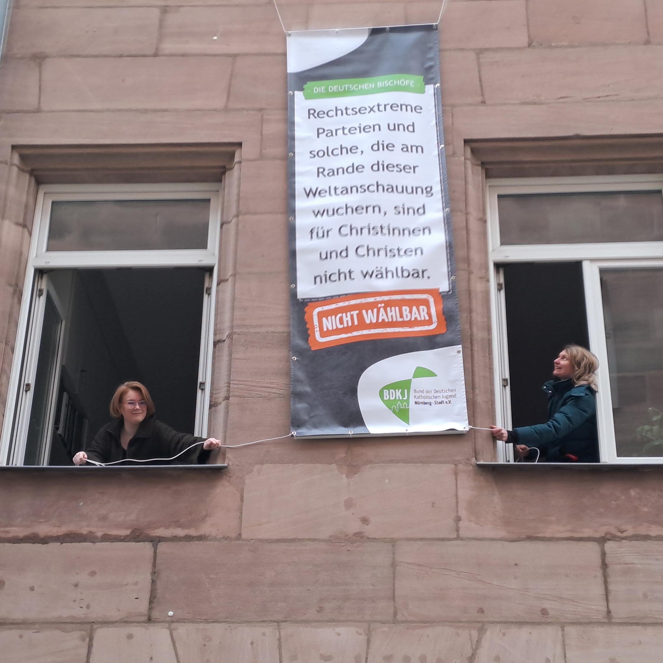 BDKJ-Referentin Sophie Will (links) und BDKJ-Stadtjugendseelsorgerin Melanie Zink hängen eines der Plakate am Haus der Katholischen Stadtkirche auf
