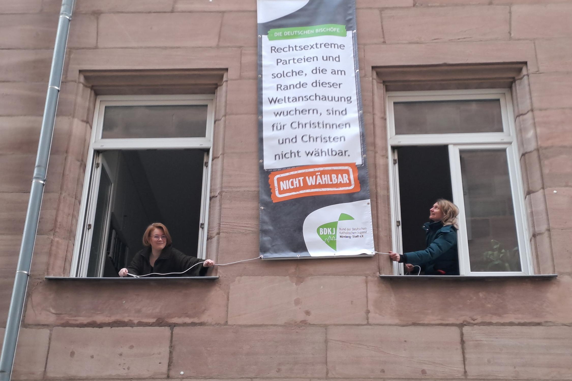 BDKJ-Referentin Sophie Will (links) und BDKJ-Stadtjugendseelsorgerin Melanie Zink hängen eines der Plakate am Haus der Katholischen Stadtkirche auf
