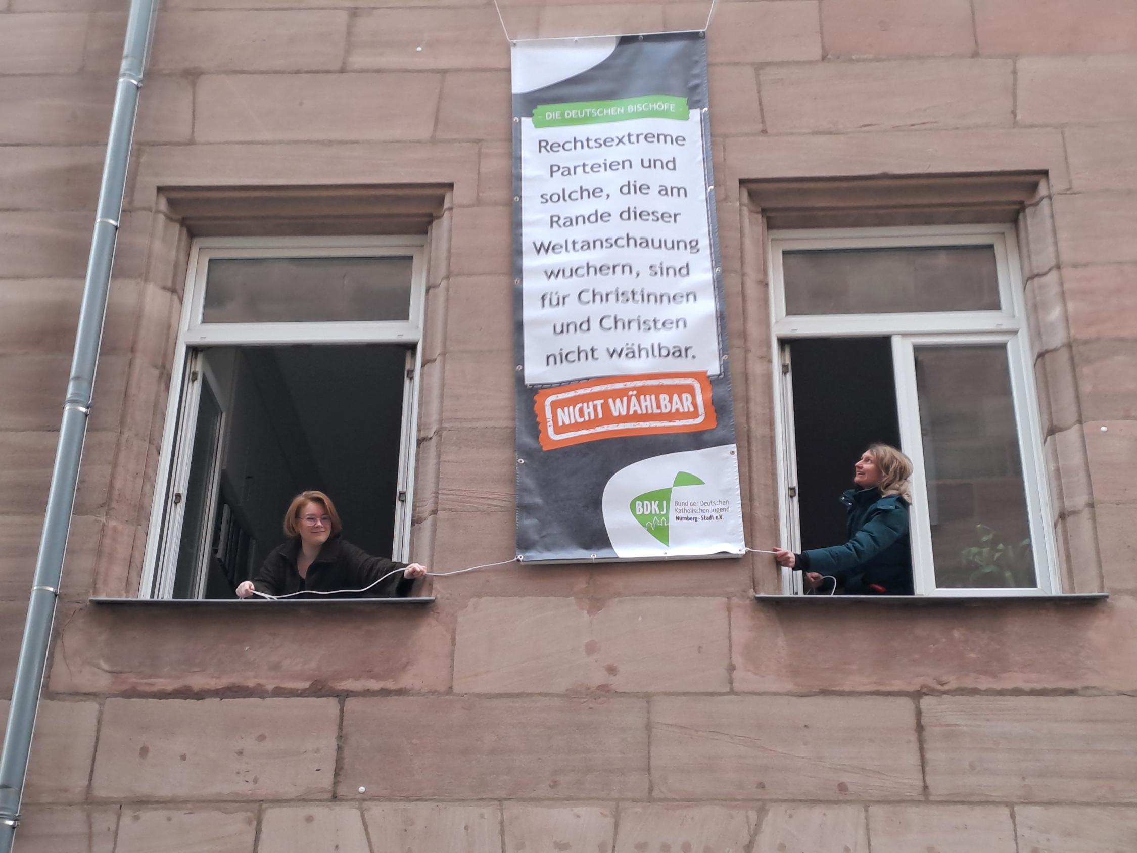 BDKJ-Referentin Sophie Will (links) und BDKJ-Stadtjugendseelsorgerin Melanie Zink hängen eines der Plakate am Haus der Katholischen Stadtkirche auf